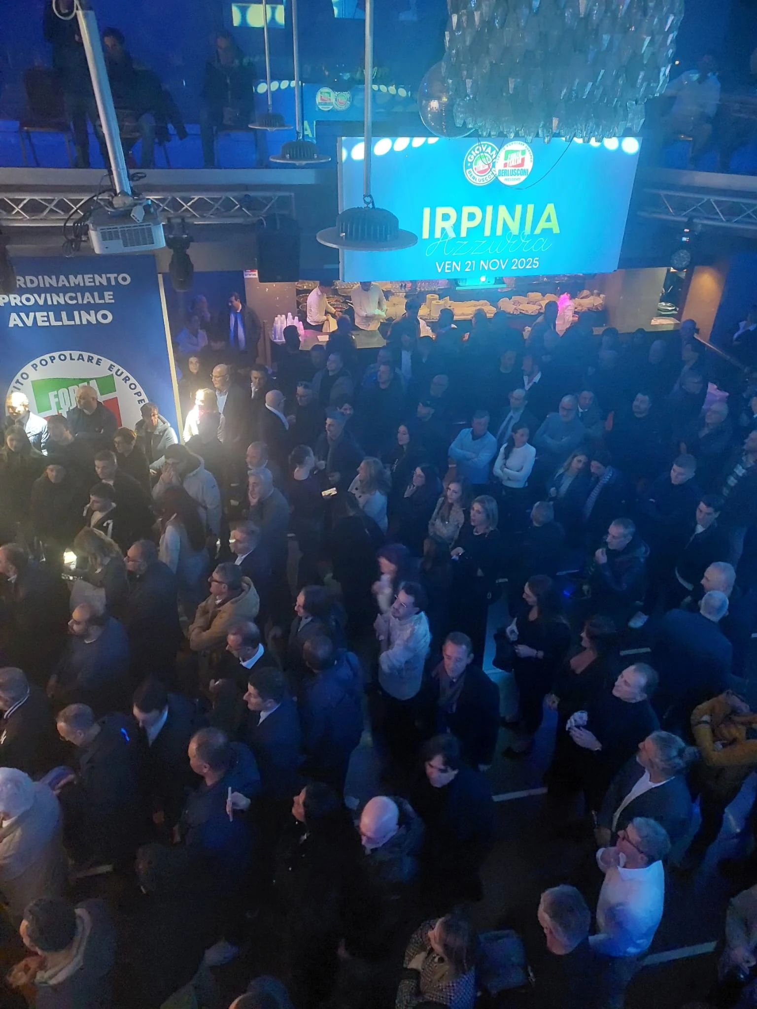 Veduta verticale di una sala affollata e del palco illuminato in blu. Archivio fotografico Regionali 2025.