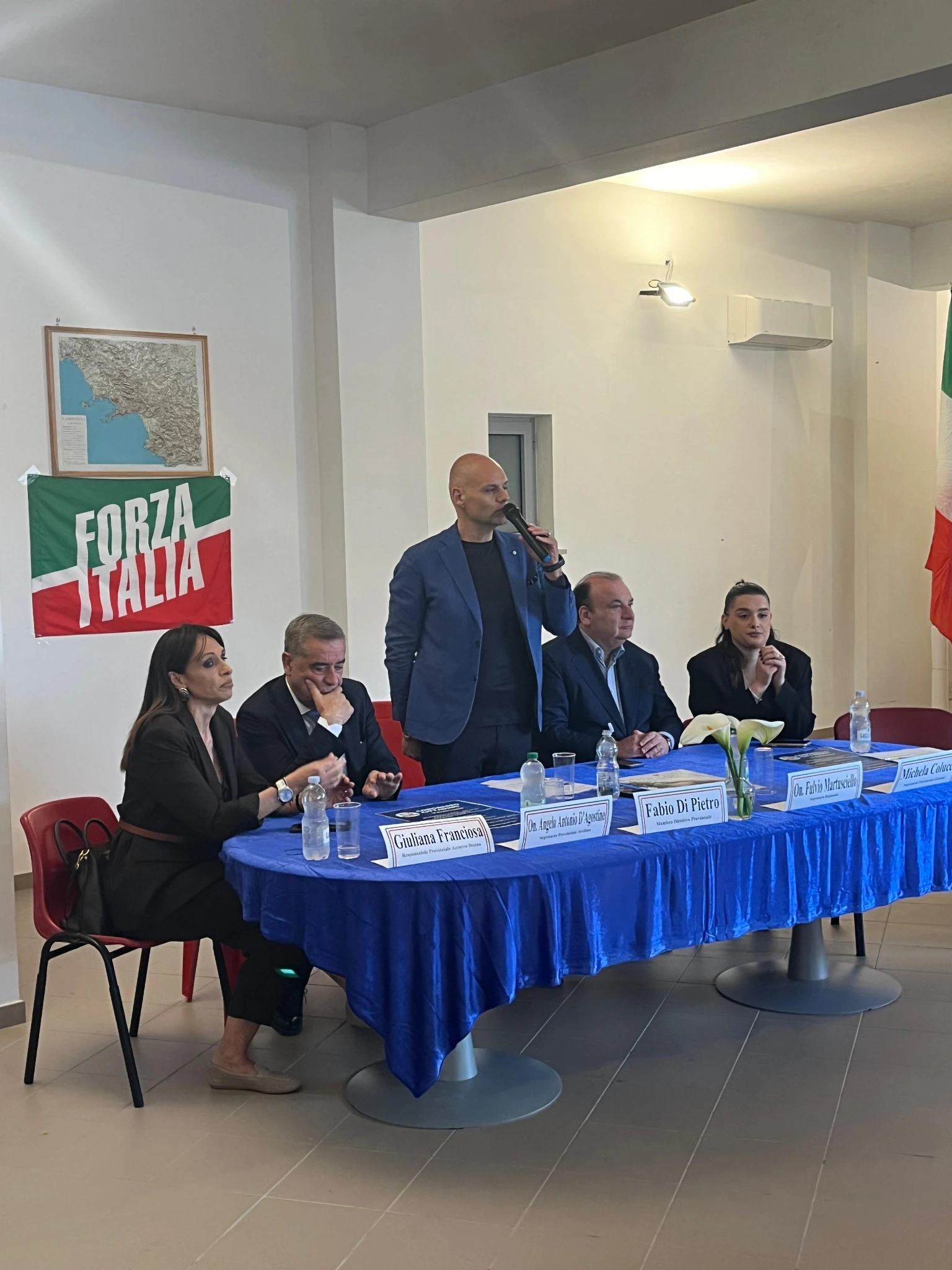 Un relatore in piedi parla al microfono, con gli altri relatori seduti al tavolo di Venticano